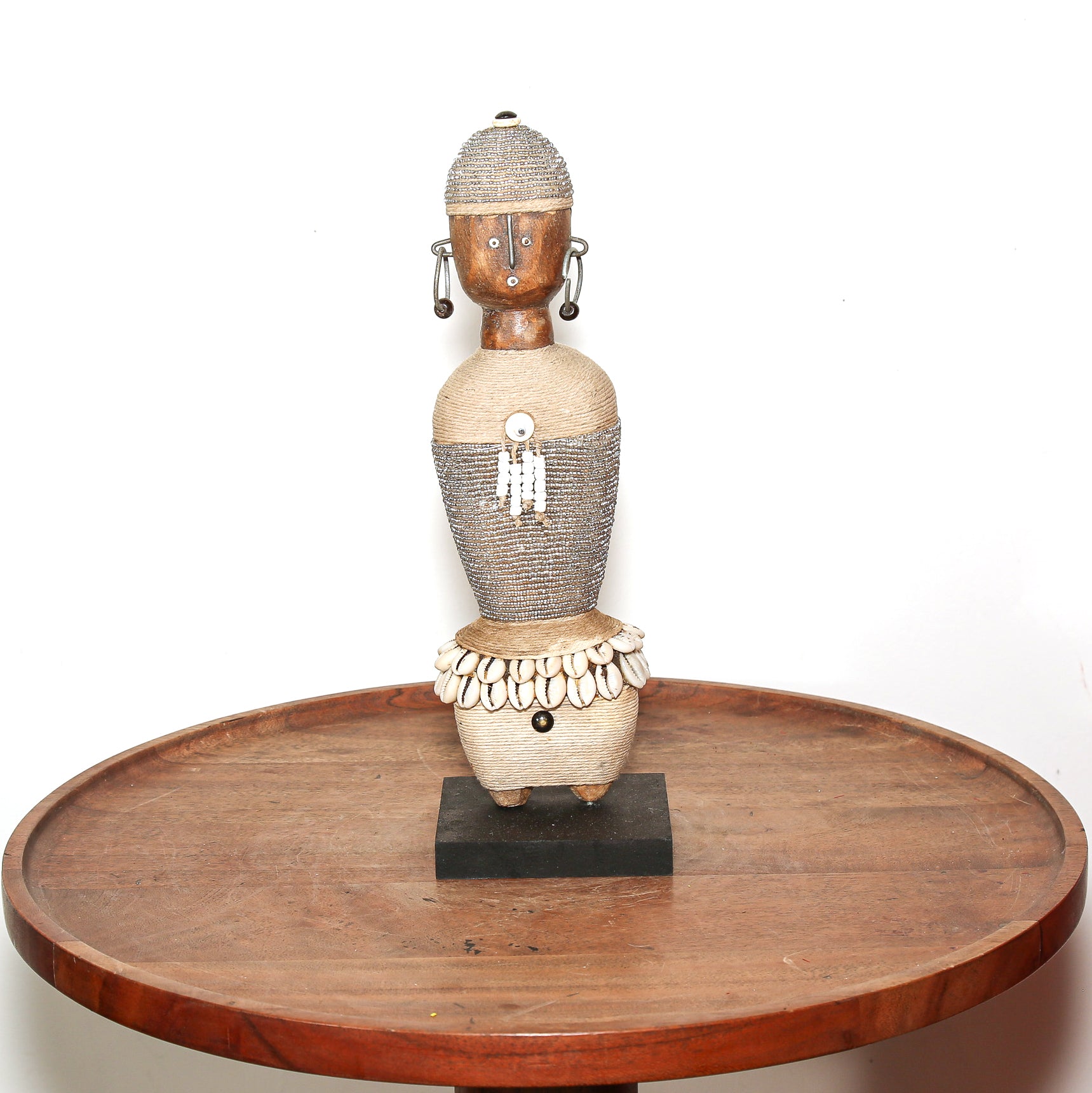 A hand-carved African wooden figurine with a textured body, decorated with white beads and shell details, displayed on a round wooden table against a white background.