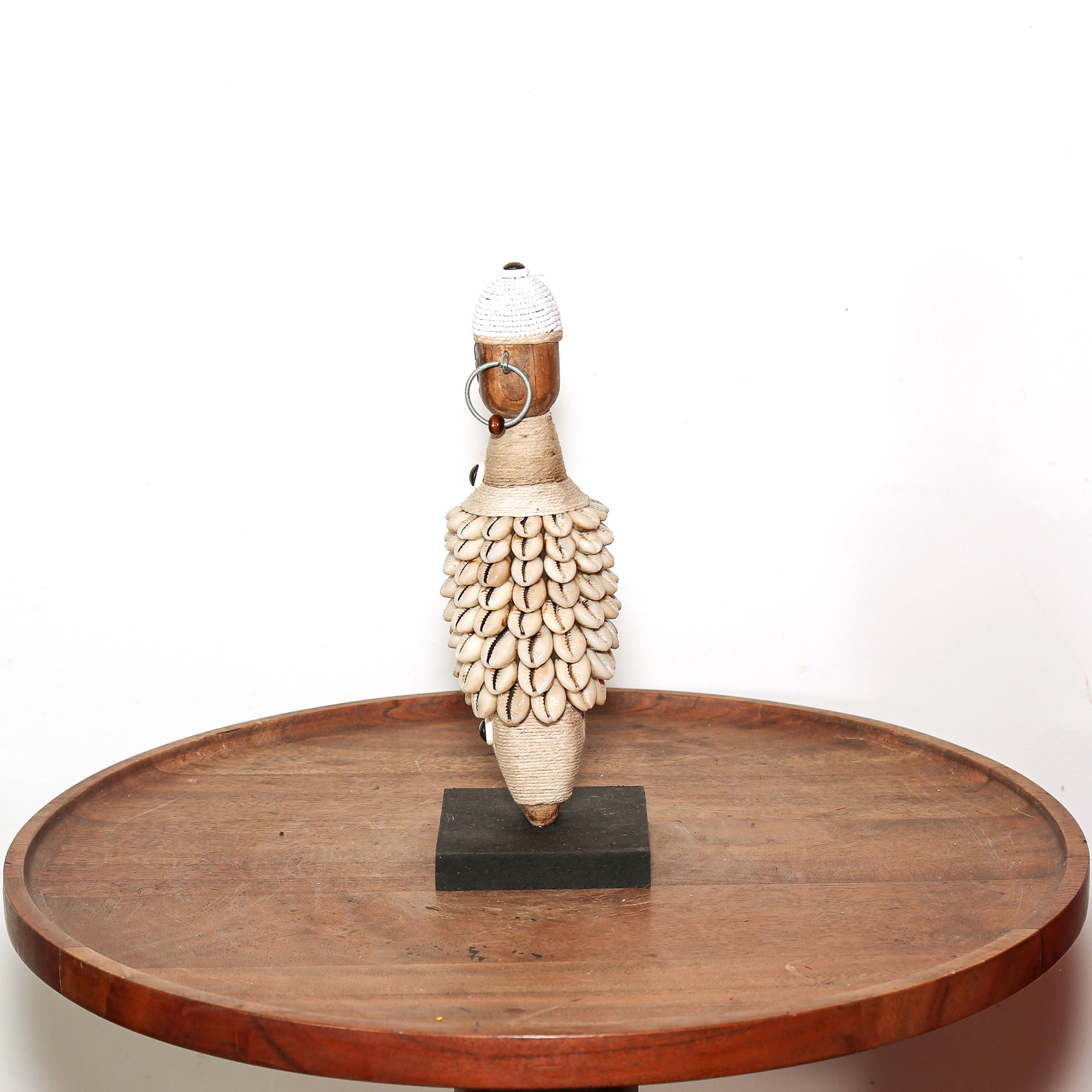 A handmade African-style wooden and shell sculpture displayed on a small black base atop a round wooden tray against a white background.