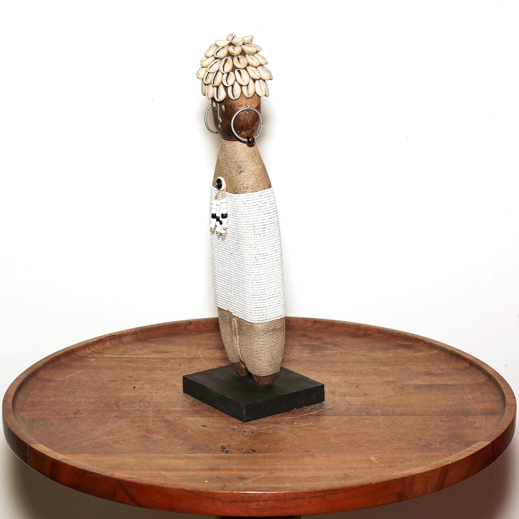 A tall African-style decorative figurine with a woven beige body, white patterned skirt, shell-like hair, and a black base, displayed on a round wooden table against a white background.