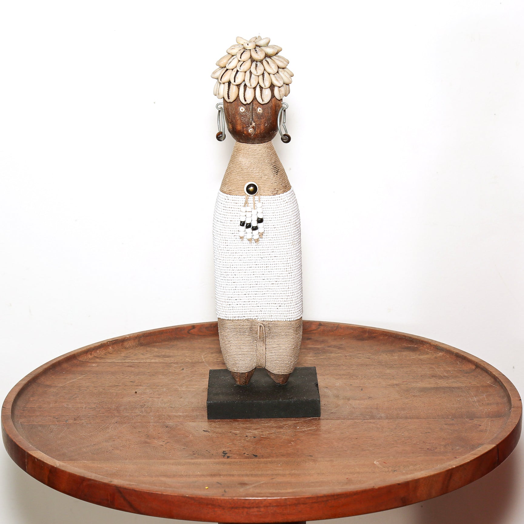 A handmade African-style wooden doll sculpture with a woven white skirt, patterned brown head, and a shell-like hairpiece, displayed on a round wooden table against a white background.