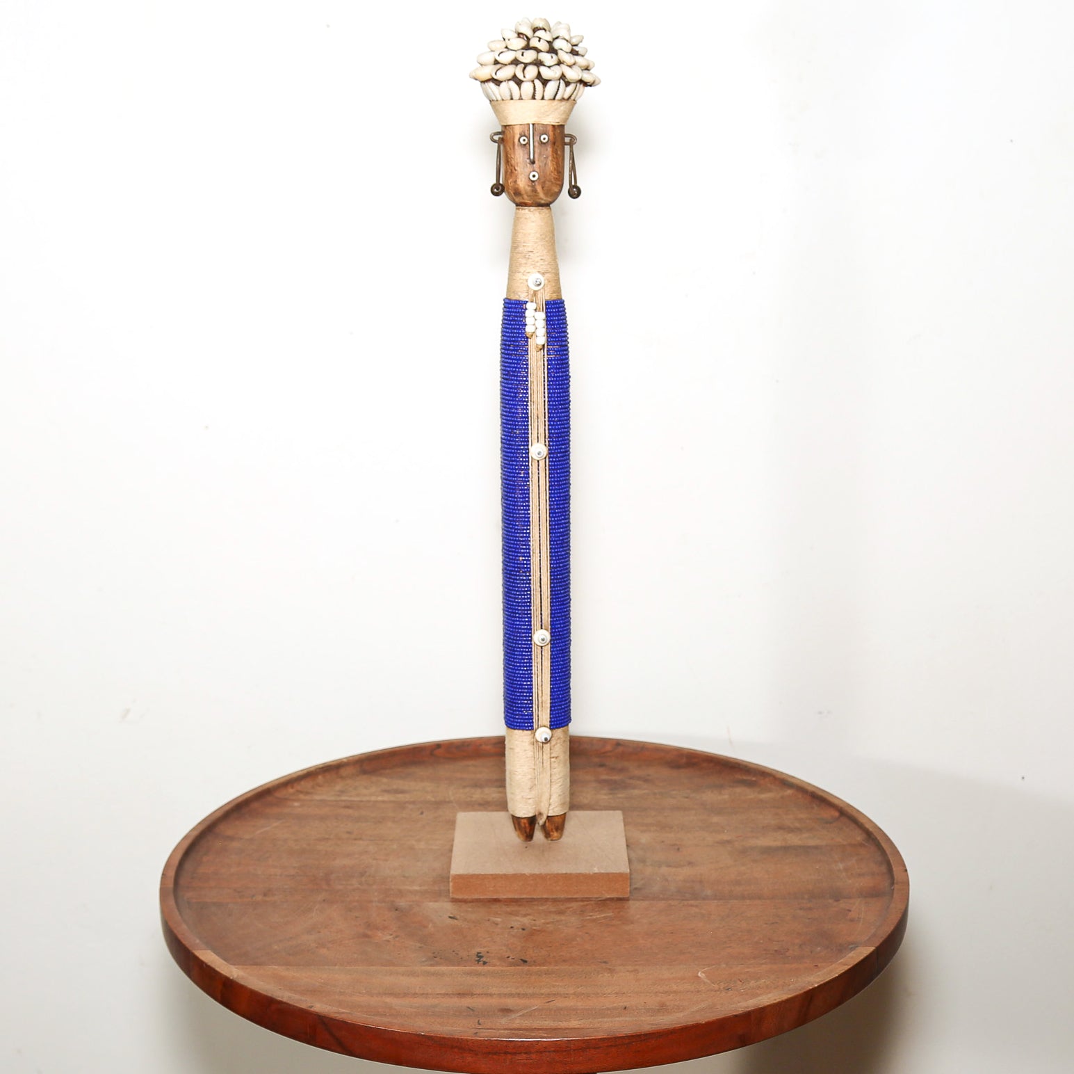 A tall handcrafted wooden sculpture with a carved face, white shell-like hair, and a bright blue wrapped body, displayed on a round wooden stand against a plain white wall.