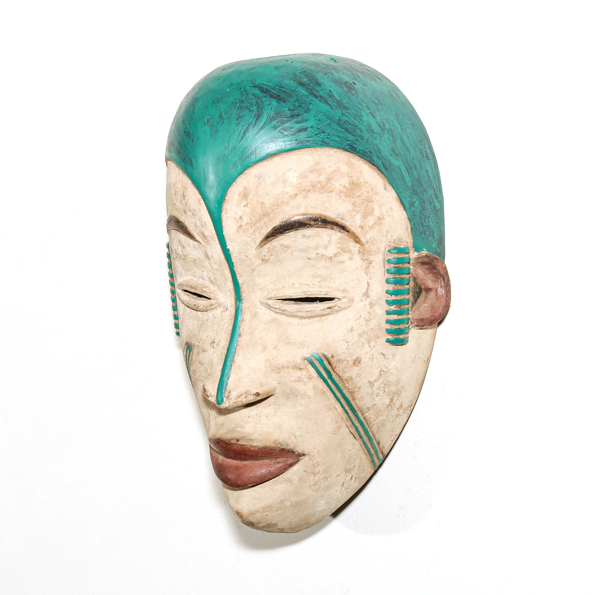 A hand-carved African wooden mask with a weathered beige face, painted teal hair, and brown lips, shown against a plain white background.