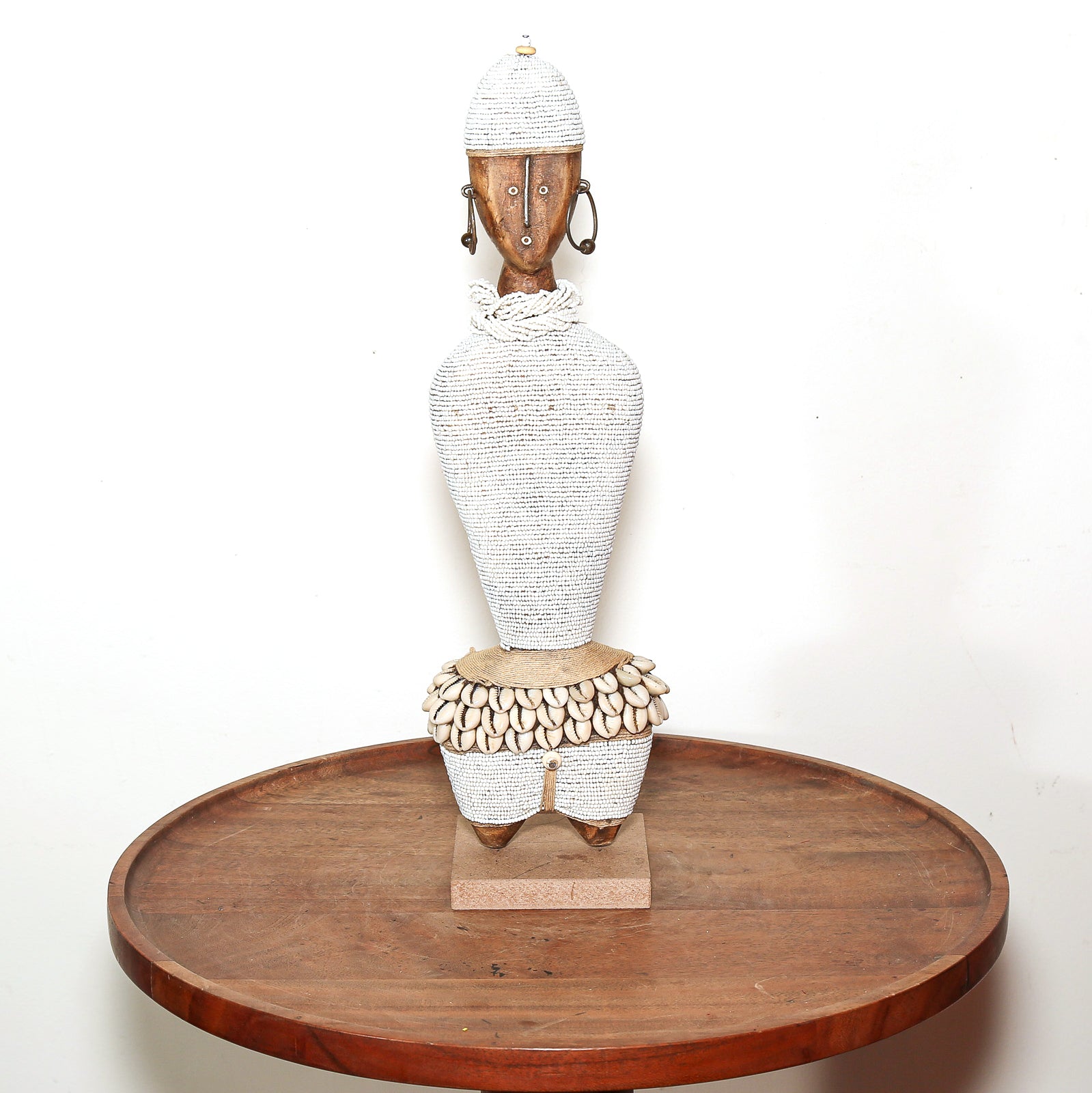 A hand-woven African-style female figurine with a textured white body, a shell-beaded waistband, and a wooden head, displayed on a round wooden table against a white background.