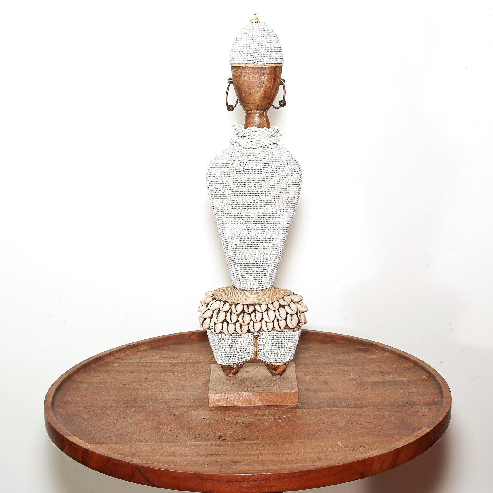 A tall African-style decorative figurine with a textured white body, a wooden head and feet, and shell-like detailing around the waist, displayed on a round wooden tray against a plain white background.