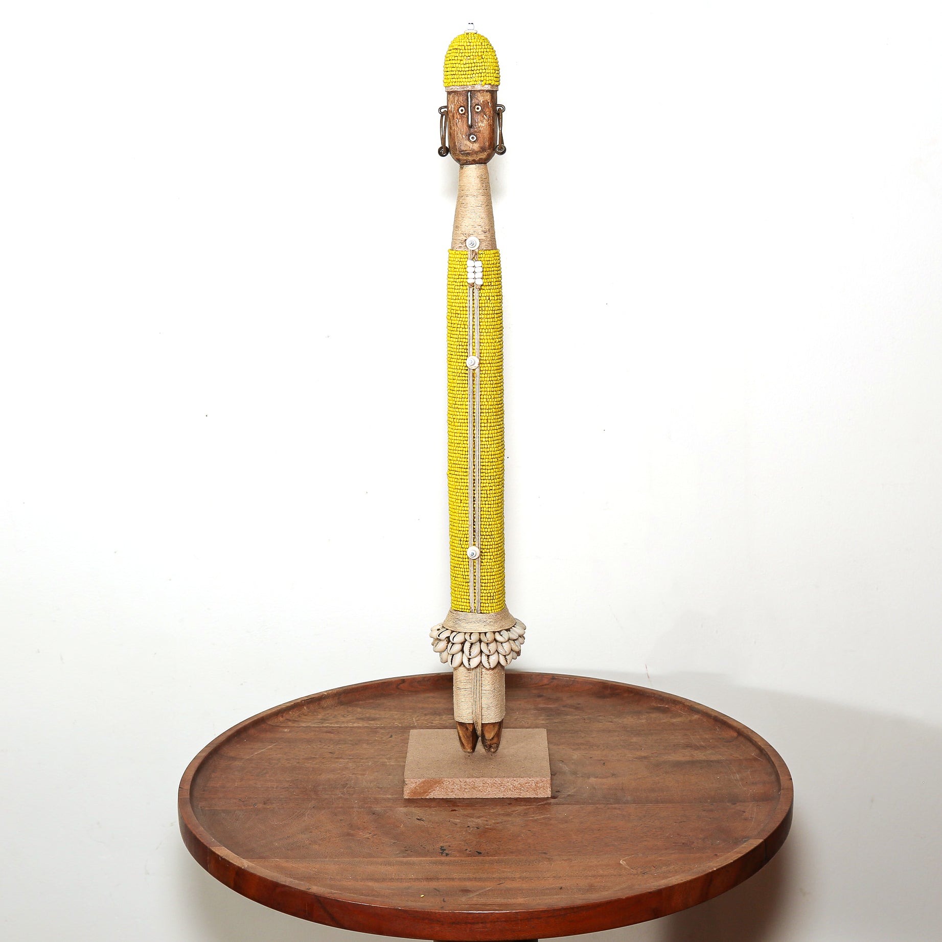 A tall hand-carved wooden sculpture of a crowned figure, decorated with a bright yellow beaded torso and a yellow headdress, displayed on a round wooden stand against a white background.