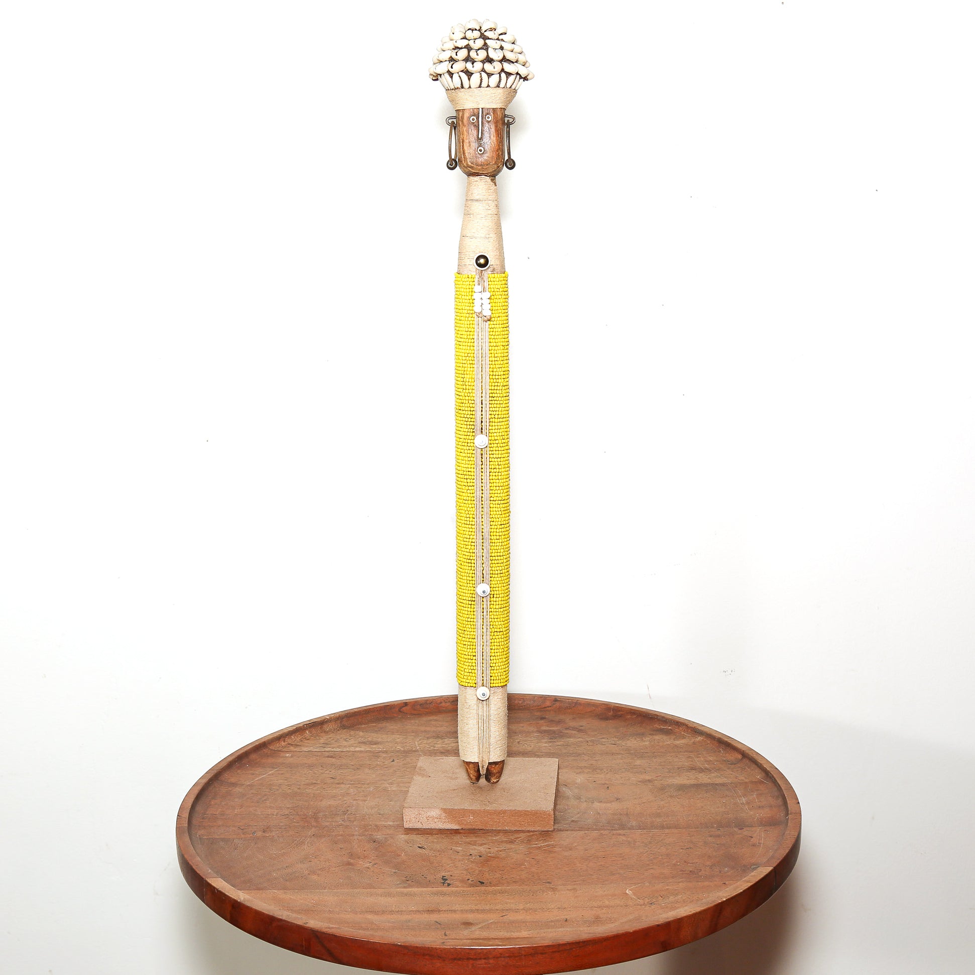 A tall decorative doll with a carved wooden top, a bright yellow beaded central section, and a small wooden base, displayed upright on a round wooden stand against a plain white background.